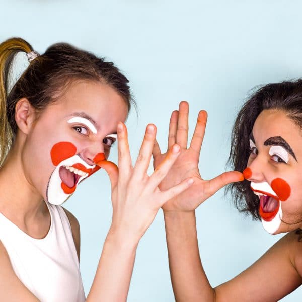 Two teenage brunettes in t-shirts and with painted faces jokingly show their long noses with their fingers palm. Isolated on gray. Family celebrates April Fool&rsquo;s Day.